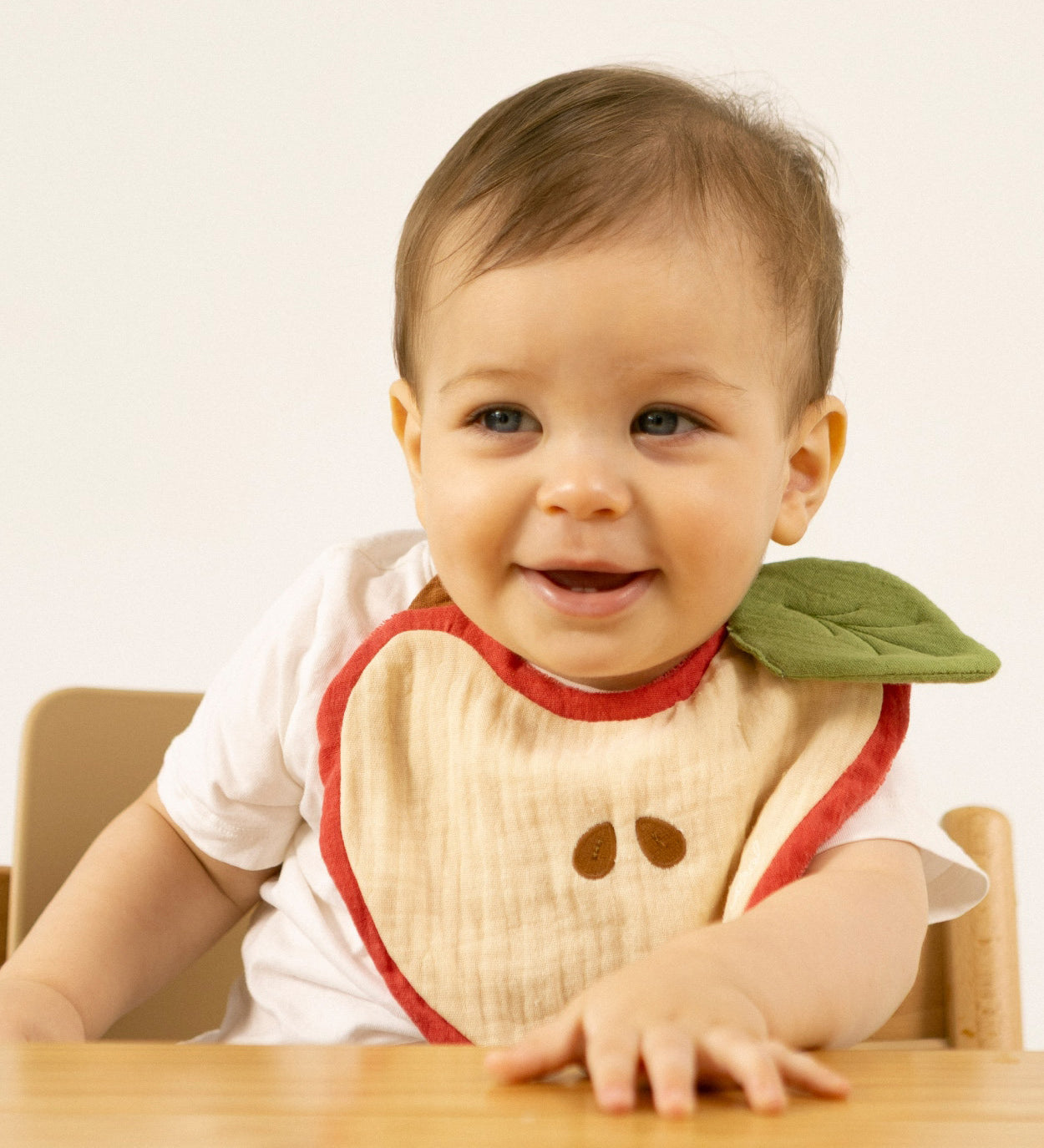 A baby sitting in a high chair and wearing the Oli & Carol Peppa the apple organic cotton baby bib available at Babipur 