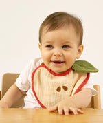 A baby sitting in a high chair and wearing the Oli & Carol Peppa the apple organic cotton baby bib available at Babipur 
