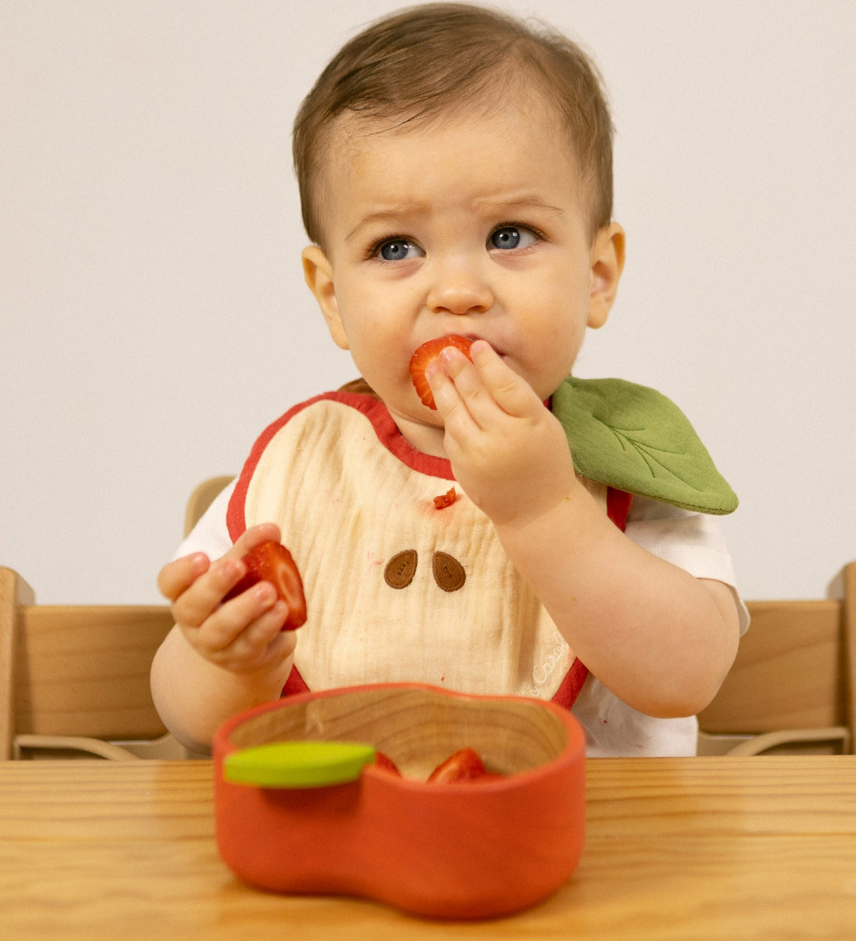 A baby sitting on a wooden high chair and  eating strawberries from the Oli & Carol Peppa the apple wooden bowl  available at Babipur. 