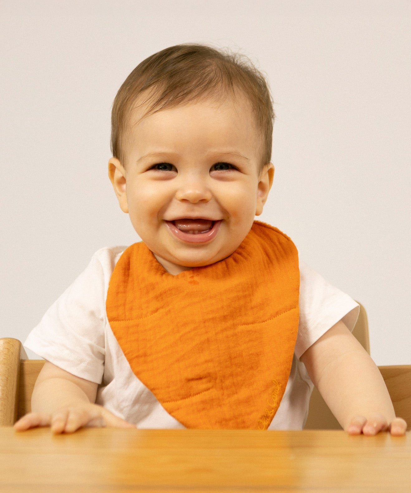 A baby sitting in a high chair and wearing the Oli & Carol Cathy the carrot organic cotton baby bib available at Babipur 