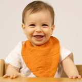 A baby sitting in a high chair and wearing the Oli & Carol Cathy the carrot organic cotton baby bib available at Babipur 