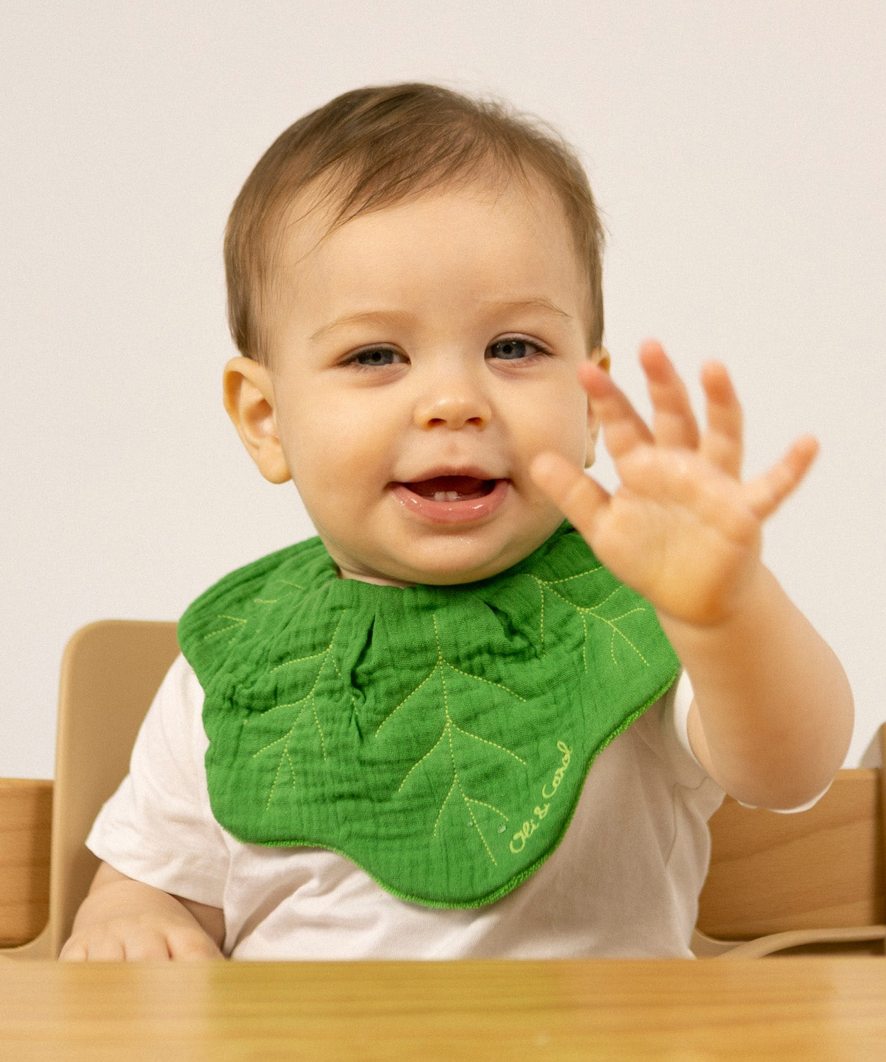 A baby sitting in a high chair and wearing the Oli & Carol Kendall the kale organic cotton baby bib available at Babipur 