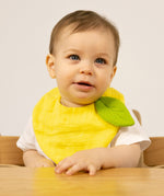 A baby sitting in a wooden high chair  and wearing the Oli & Carol Lou the lemon baby bib available at Babipur. 