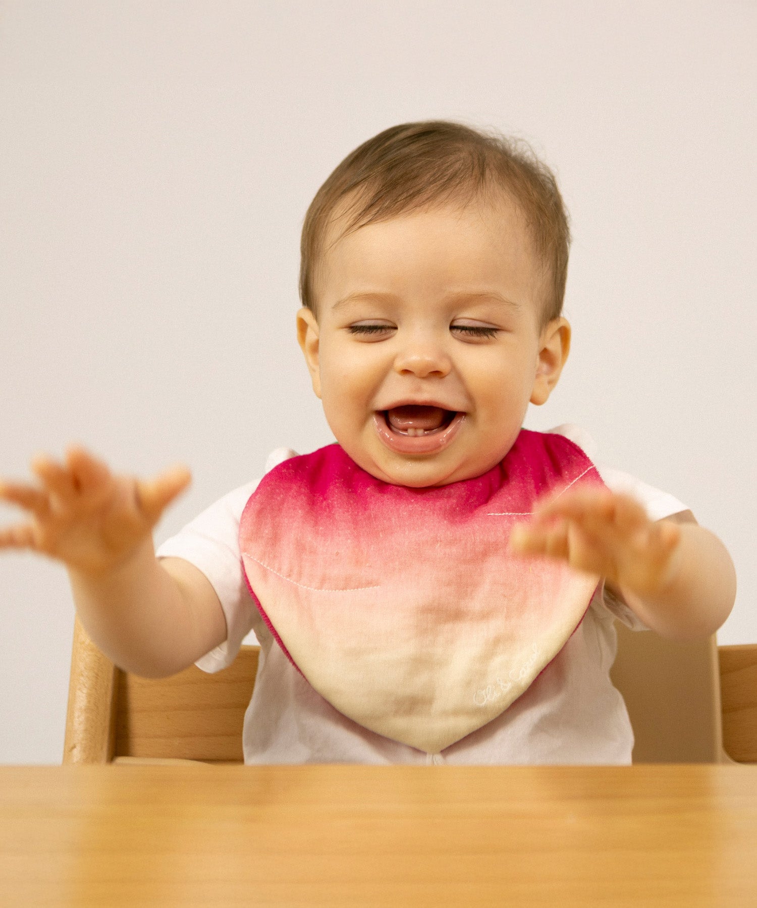 A baby sitting in a high chair and wearing the Oli & Carol Ramona the radish organic cotton baby bib available at Babipur 