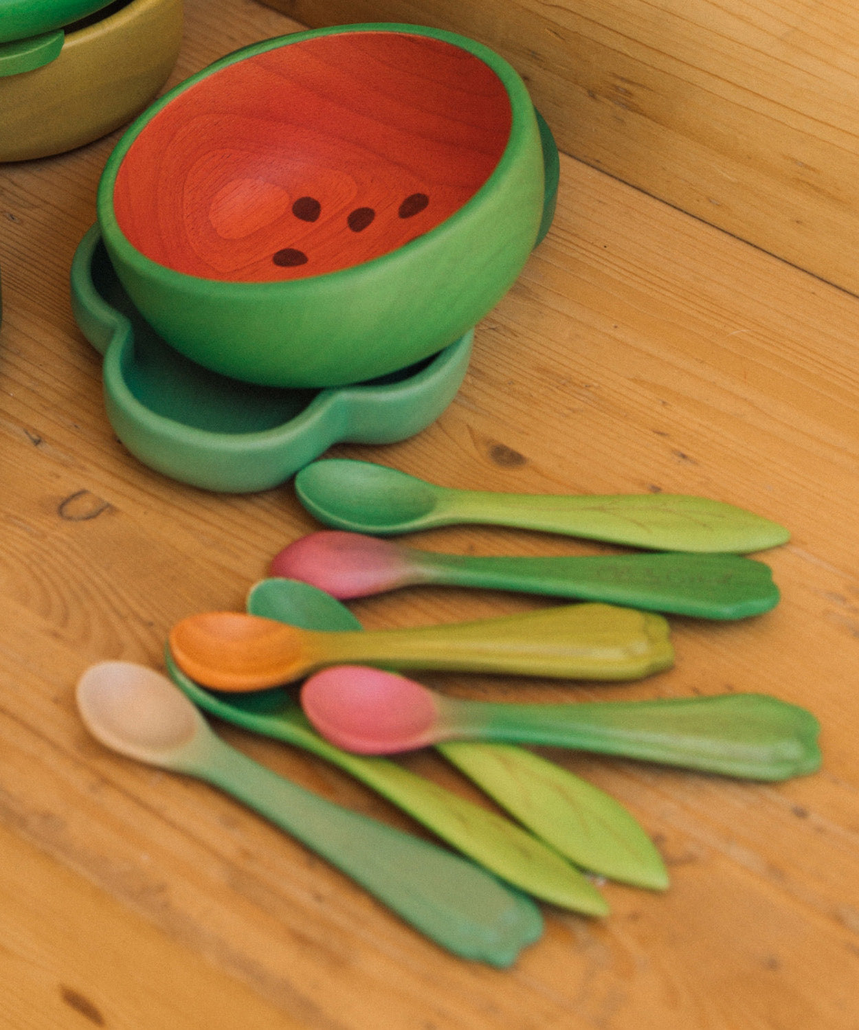 OIi & Carol wooden baby weaning spoons on a wooden surface next to a watermelon bowl. Both 2 packs are available at Babipur.