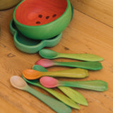 OIi & Carol wooden baby weaning spoons on a wooden surface next to a watermelon bowl. Both 2 packs are available at Babipur.