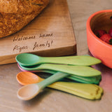 A stack of Oli & Carol wooden weaning baby spoons on a wooden surface showing the leek and kale design and the carrot and radish design. Both 2 packs are available at Babipur.