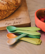 A stack of Oli & Carol wooden weaning baby spoons on a wooden surface showing the leek and kale design and the carrot and radish design. Both 2 packs are available at Babipur.
