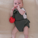 A baby laying down on a cream blanket, playing with the Oli & Carol Pepa Apple Pacifier Holder and Teething Toy
