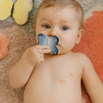 A child holding the Oli & Carol Natural Rubber Mini Teether - Blues The Butterfly to their mouth, and laying on a colourful rug