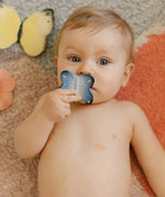 A child holding the Oli & Carol Natural Rubber Mini Teether - Blues The Butterfly to their mouth, and laying on a colourful rug
