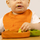A baby sitting in a wooden high chair eating and wearing the Oli & Carol Cathy the carrot baby bib available at Babipur. 