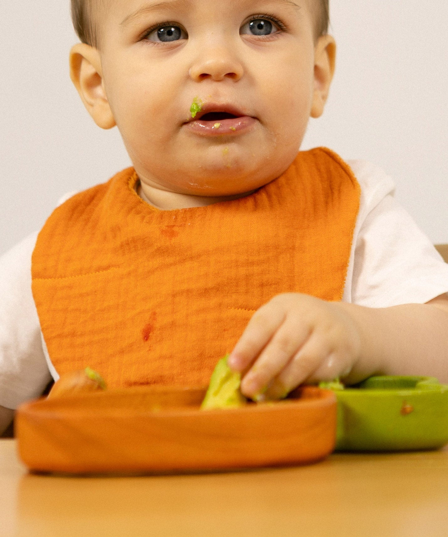 A baby sitting in a wooden high chair eating and wearing the Oli & Carol Cathy the carrot baby bib available at Babipur. 