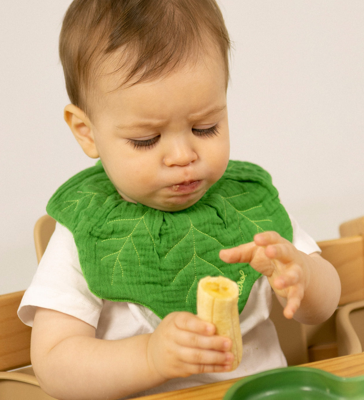 A baby sitting in a wooden high chair eating a banana and wearing the Oli & Carol Kendall the kale baby bib available at Babipur. 