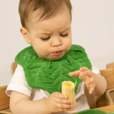 A baby sitting in a wooden high chair eating a banana and wearing the Oli & Carol Kendall the kale baby bib available at Babipur. 