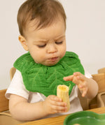 A baby sitting in a wooden high chair eating a banana and wearing the Oli & Carol Kendall the kale baby bib available at Babipur. 