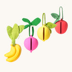 The Oil & Carol Fruit & Veggie Pacifier Cases, made from Natural Rubber in a diagonal row on a cream background. There is a yellow banana, a pink/purple radish, a yellow lemon and a red apple. The image also showing the slot where the pacifier/dummy fits into the holder case