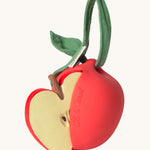 The Pepa The Apple Pacifier Holder on a cream background. The image shows an Oli & Carol apple teething toy half way inside the holder. The apple is smooth and has a green muslin leaf attachment on the top to tie the holder to a pram handle, high chair etc.