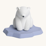 Oli & Carol Polar Bear Bath Toy. A bright white polar bear, natural rubber bath toy is sitting on a flat, light blue/lilac iceberg on a cream background. The polar bear is white, has a slightly textured body and has a black nose. 