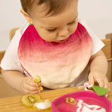 A baby eating fruit and wearing the Oli & Carol Ramona the radish organic cotton baby bib available at Babipur 