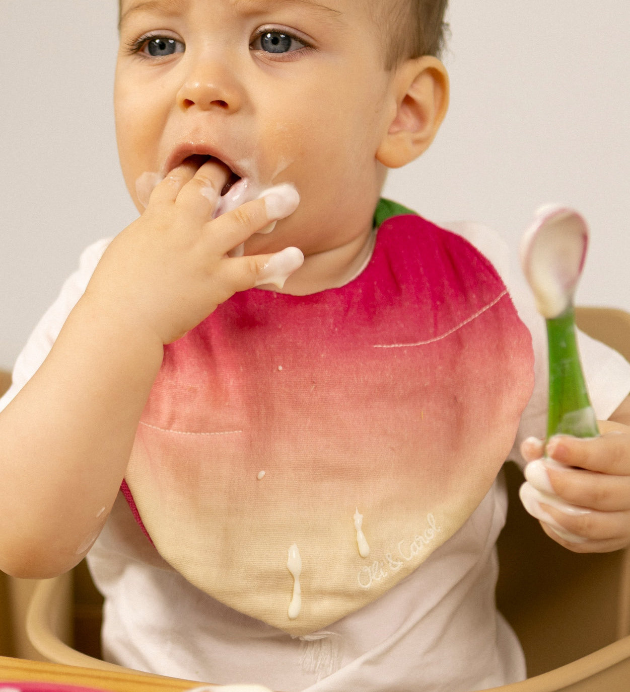 A baby sitting in a wooden high chair eating yogurt and wearing the Oli & Carol Ramona the radish baby bib available at Babipur. 