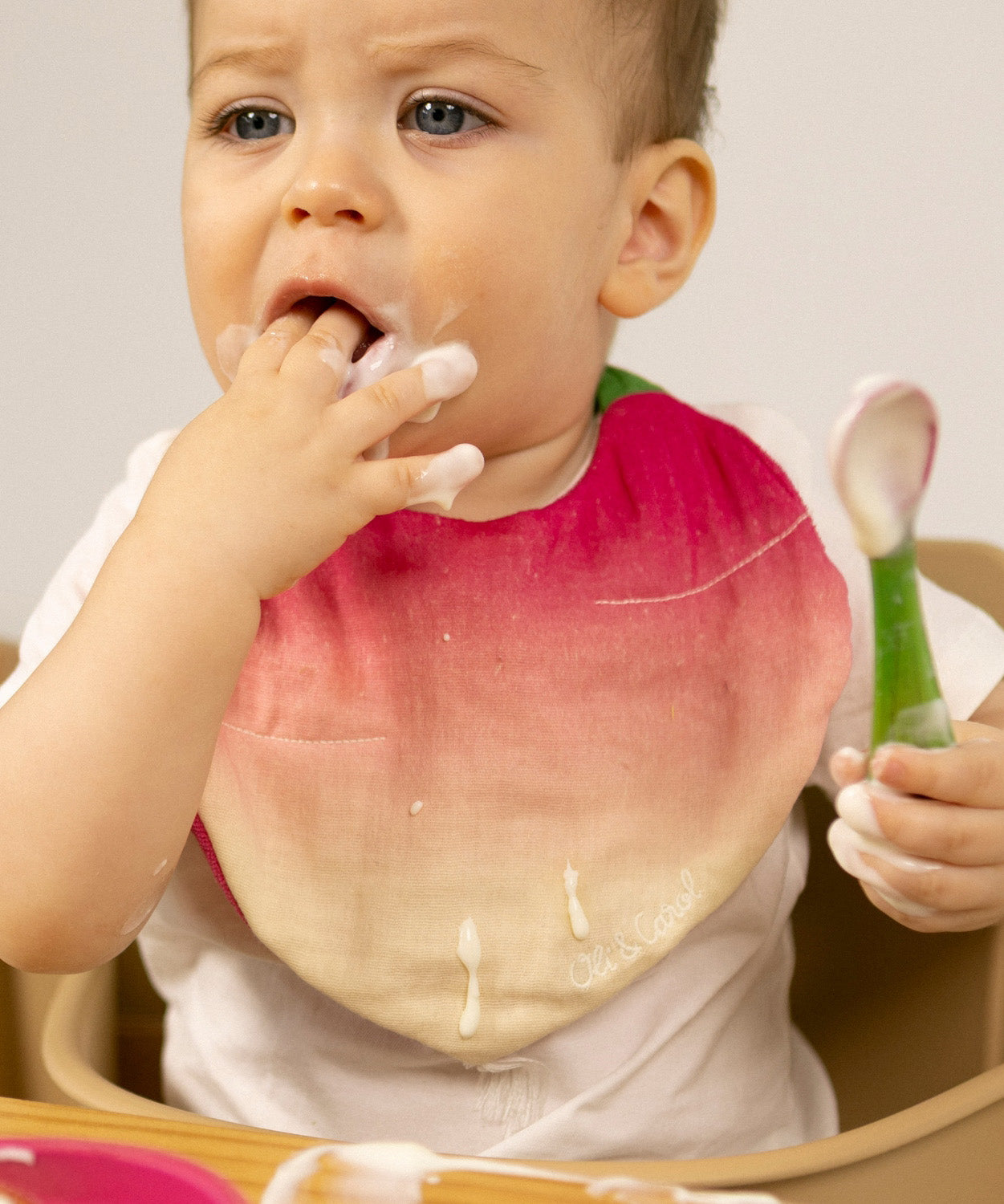 A baby sitting in a wooden high chair eating yogurt and wearing the Oli & Carol Ramona the radish baby bib available at Babipur. 