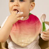 A baby sitting in a wooden high chair eating yogurt and wearing the Oli & Carol Ramona the radish baby bib available at Babipur. 