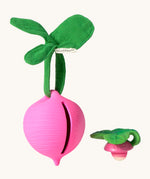 The Ramona Radish Pacifier Holder on a cream background. The image shows an Oli & Carol radish teething toy next to the holder, and the slot where the pacifier or teether can go inside the holder. The radish is textured and has a green muslin leaf attachment on the top to tie the holder to a pram handle, high chair etc.