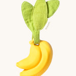 The Ana Banana Pacifier Holder on a cream background. The banana is smooth and has a green muslin leaf attachment on the top to tie the holder to a pram handle, high chair etc.