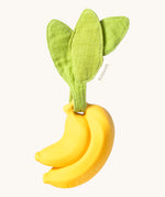 The Ana Banana Pacifier Holder on a cream background. The banana is smooth and has a green muslin leaf attachment on the top to tie the holder to a pram handle, high chair etc.