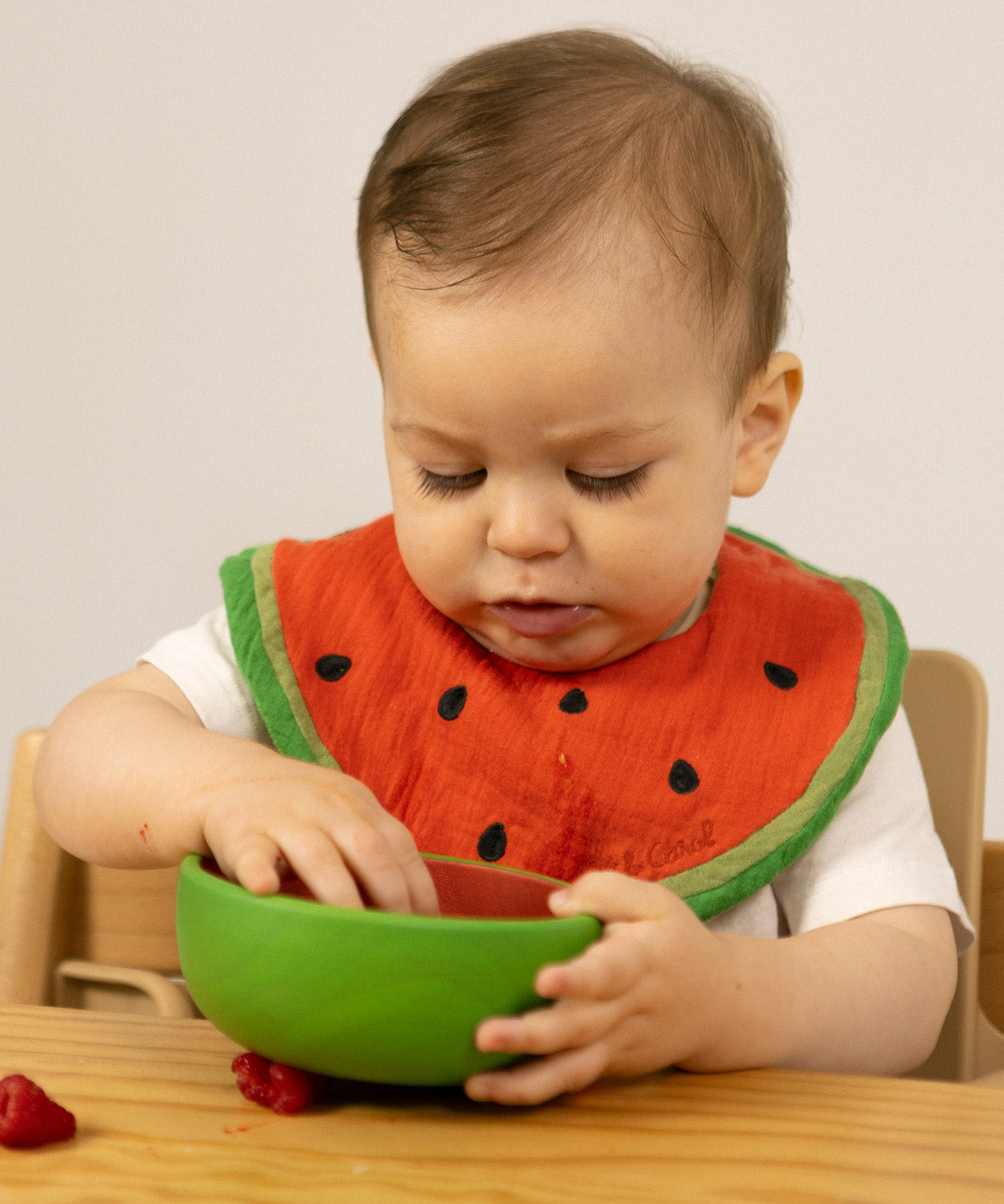 A baby sitting in a wooden high chair eating fruit and wearing the Oli & Carol Wally the watermelon baby bib available at Babipur. 