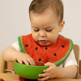 A baby sitting in a wooden high chair eating fruit and wearing the Oli & Carol Wally the watermelon baby bib available at Babipur. 