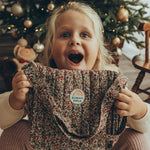 A child looking very excited to have received the Olli Ella Carrie Convertible Changing Set - Garland. The child is holding the toy changing bag in their hands, with a surprised and happy expression on their face.
