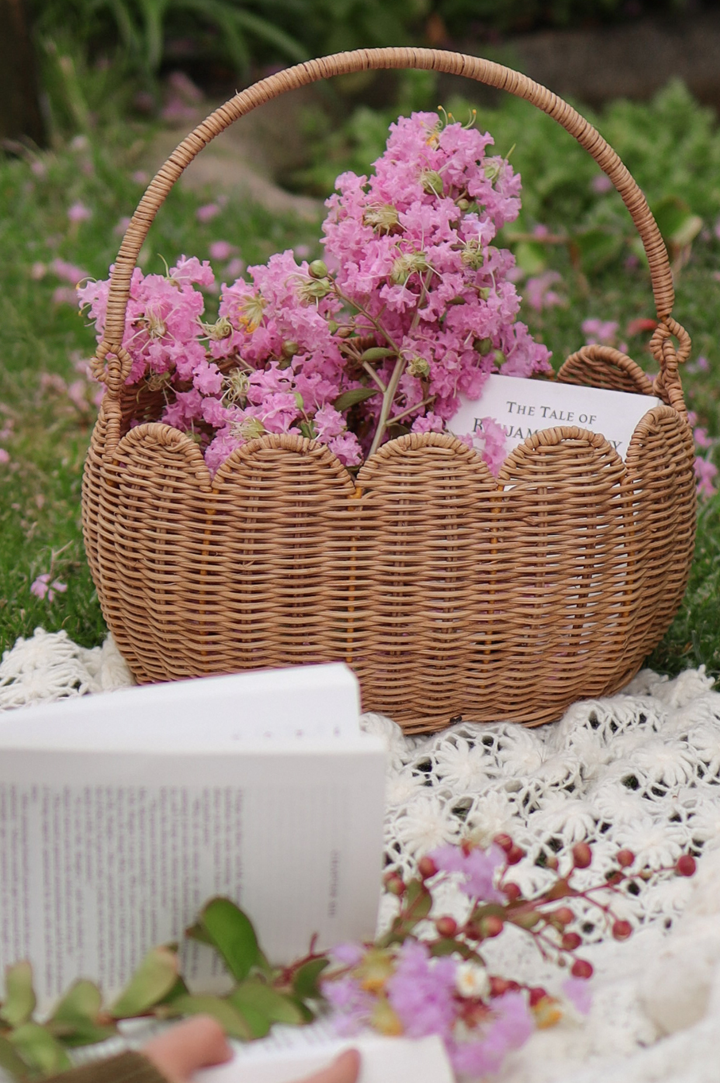 Olli Ella Rattan Petal Basket in brown with scalloped edges and a handle filled with purple flowers to represent Olli Ella Adult Baskets and Bags at Babipur.