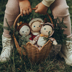 A child is holding the Olli Ella Rattan Poinsettia basket by the handle, in their hands. The basket is on a grassy floor with Christmas Dinky Dinkums inside, and small pine tree branches to give a festive feel.