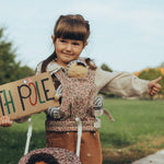 A child holding a cardboard sign saying "North Pole" in colourful letters whilst wearing an Ollie Ella Dinkum Doll in the Olli Ella Dinkum Dolls Carrier - Garland. There is also the top part of an Olli Ella Stroller by the childs legs, with a Dinkum Doll sat inside.