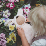 A little girl holding a Olli Ella cozy dinkum bunny pip doll surrounded by flowers. This is a soft and fluffy doll with a sleeping face, cute floppy ears and available at Babipur.