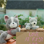 A grey and white gizmo Olli Ella dinkum dog standing next to banjo the puppy that's lying on a Olli Ella natural basket.