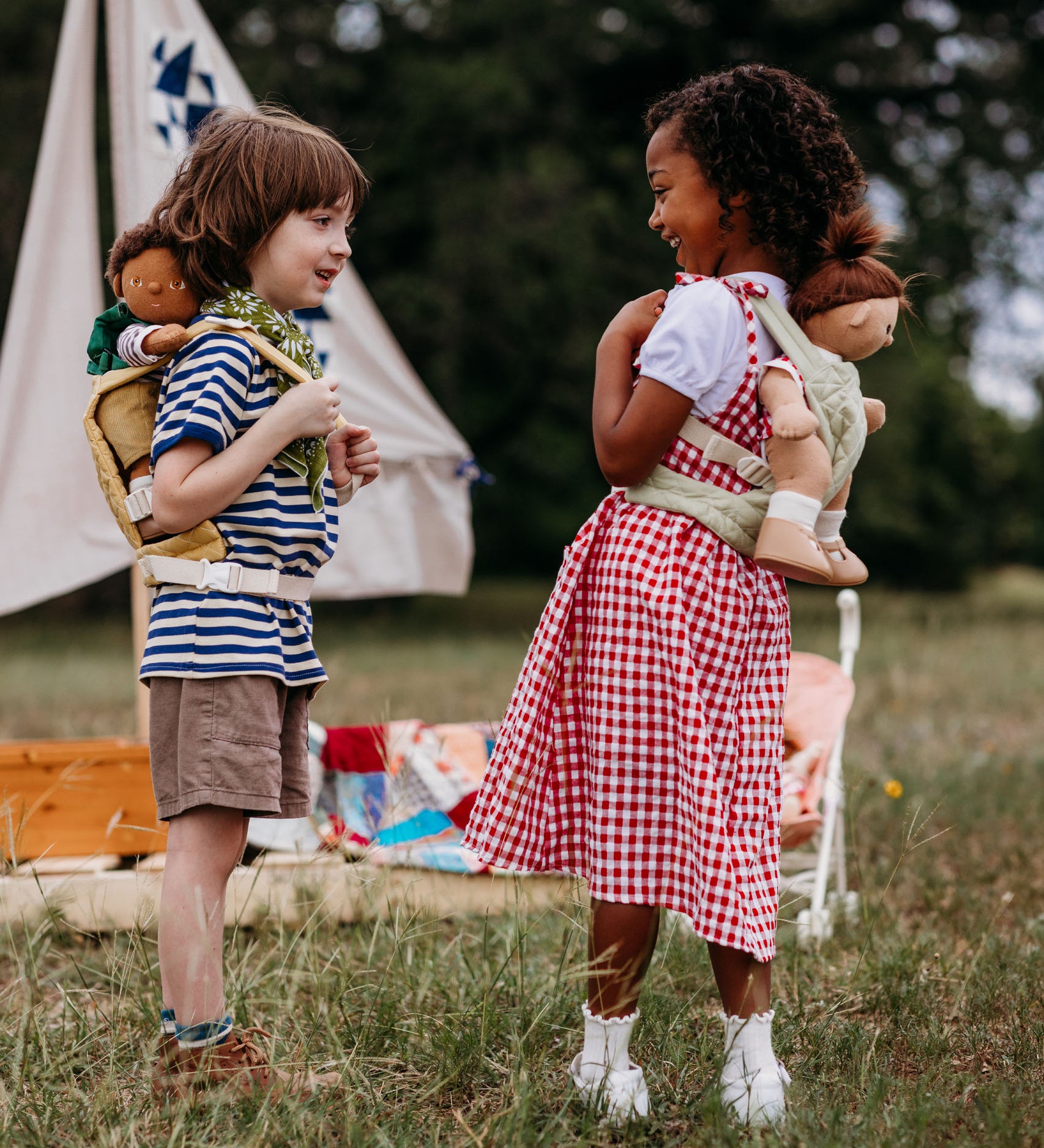 2 Children playing outside and wearing the Olli Ella dinkum doll quilted carriers in sage and honey colours with dinkum dolls sitting inside. These toy carriers are available at Babipur.