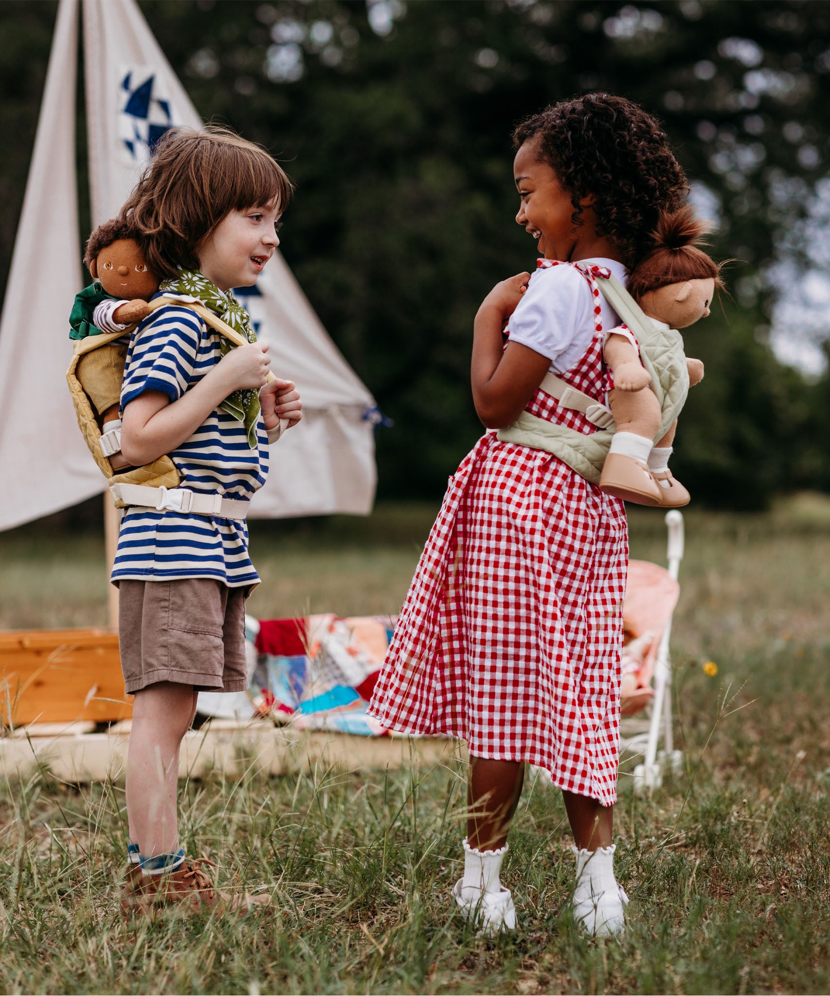 2 Children playing outside and wearing the Olli Ella dinkum doll quilted carriers in sage and honey colours with dinkum dolls sitting inside. These toy carriers are available at Babipur.