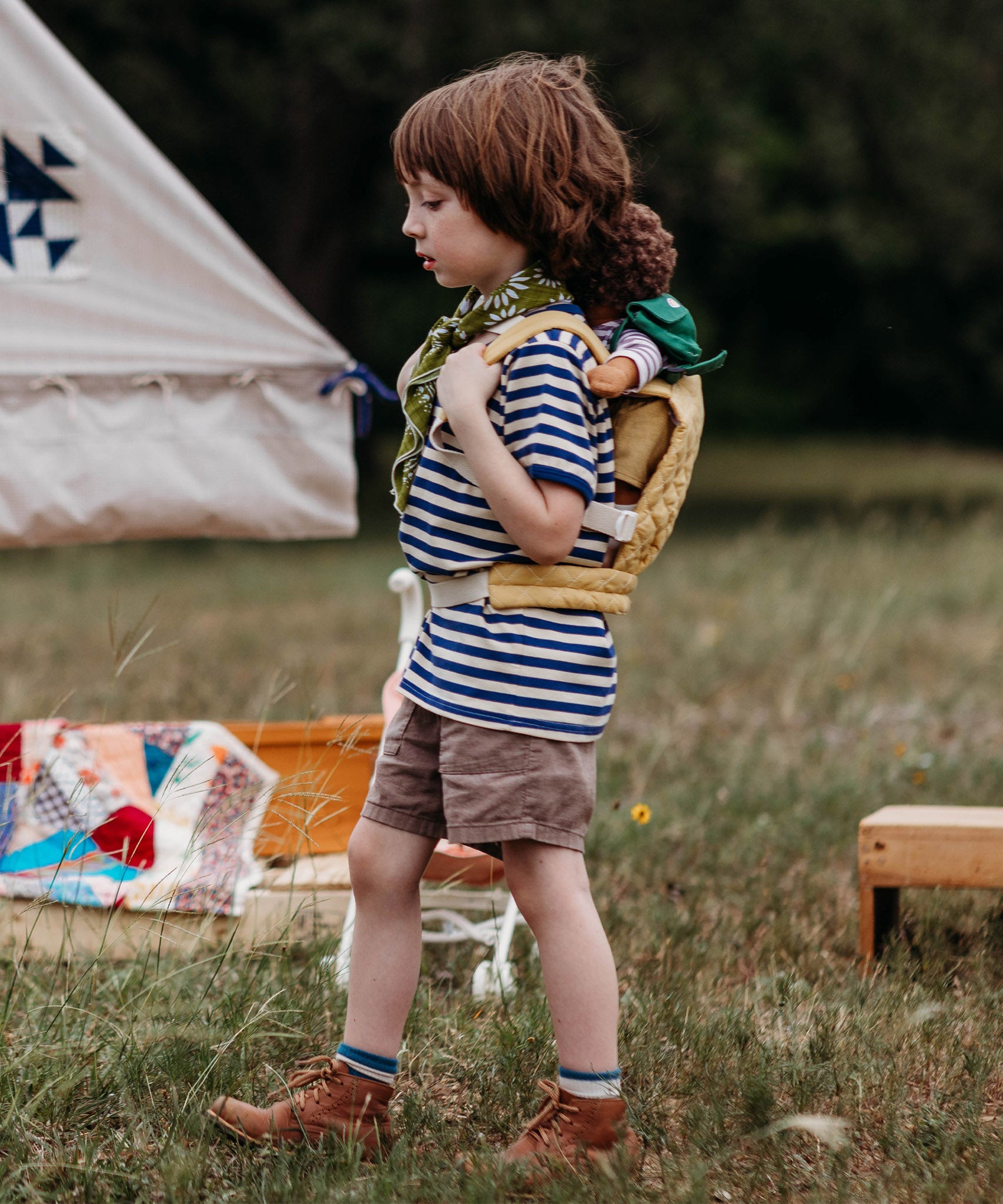 A child walking outside and wearing the Olli Ella dinkum doll quilted carrier in  honey colour with a dinkum doll sitting on it's back. This toy carriers is available at Babipur.