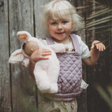 A child holding a Olli Ella cozy bunny doll whilst wearing the Olli Ella quilted toy carrier in lavender. This toy carrier is available at Babipur.