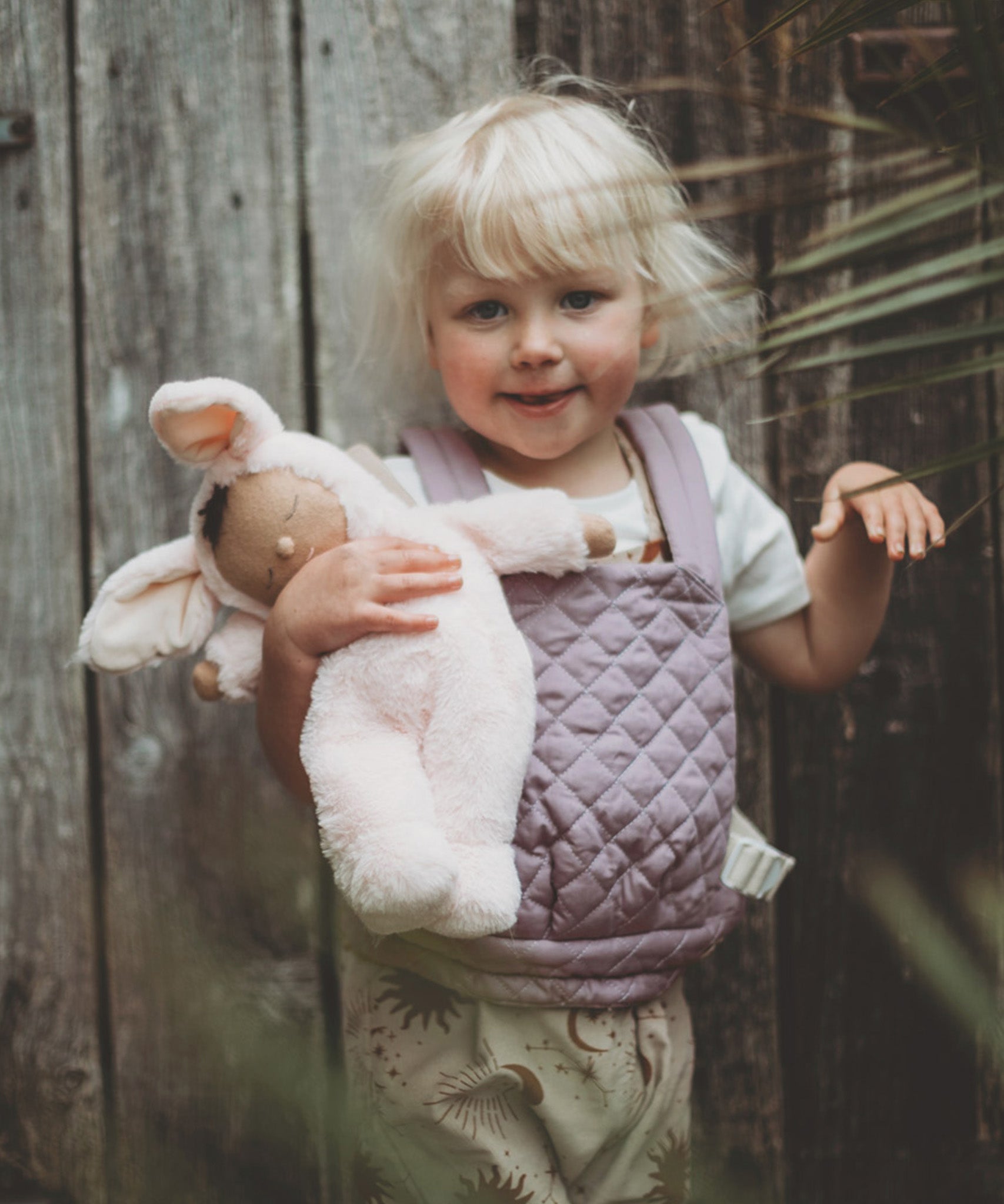 A child holding a Olli Ella cozy bunny doll whilst wearing the Olli Ella quilted toy carrier in lavender. This toy carrier is available at Babipur.