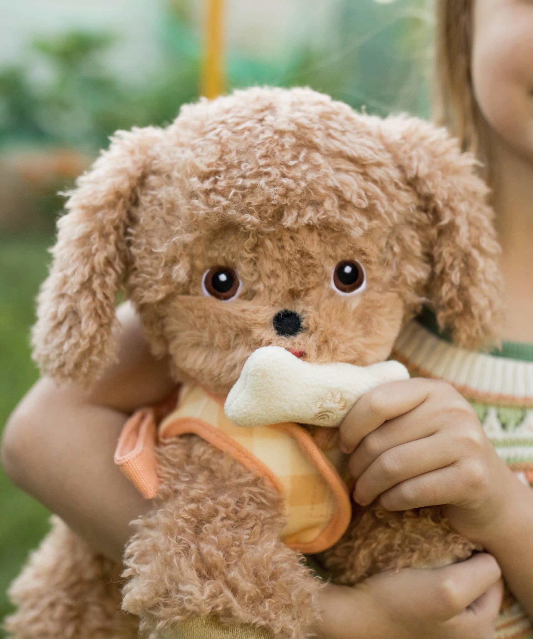 A child holding the soft and fluffy brown Olli Ella Goldie dinkum dog with a magnetic bone in its mouth.