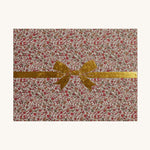 The packaging for the Olli Ella Dinkum Dolls Carrier in Garland. The packaging has the same colourful, vintage, floral print and has a gold painted bow on top. The image is on a cream background