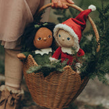 A child is carrying their Festive Jolly Dolly Dinkum Dolls, Paige Pudding and Sammie Santa, in a natural Ollie Ella Poinsettia Carry Basket. Inside the basket with the dolls are branches from a pine tree, to give a festive feel.
