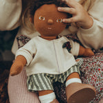 A child sat with an OllI Ella Dinkum Doll on their knee, which is wearing the Olli Ella Dinkum Doll Holiday Outfit Set.