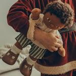 A child wearing a Santa outfit, is holding a Olli Ella Dinkum Doll under their arm, which is wearing the Olli Ella Dinkum Doll Holiday Outfit Set