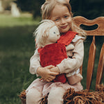 A child is sat down on a antique wooden chair, and is carrying a Dinkum Doll in the Olli Ella Dinkum Dolls Petal Carrier - Red Velvet. The child is happy and is cuddling the doll. The chair has a brown fluffy rug on the seat.