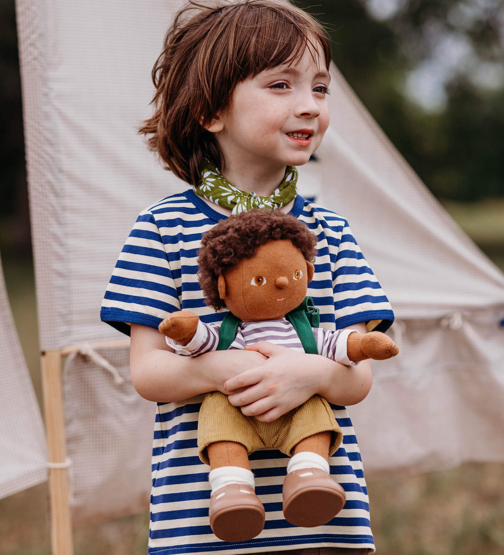 A child playing outside and holding a dinkum doll that's wearing the Olli Ella rambler dinkum doll outfit set available at Babipur. 